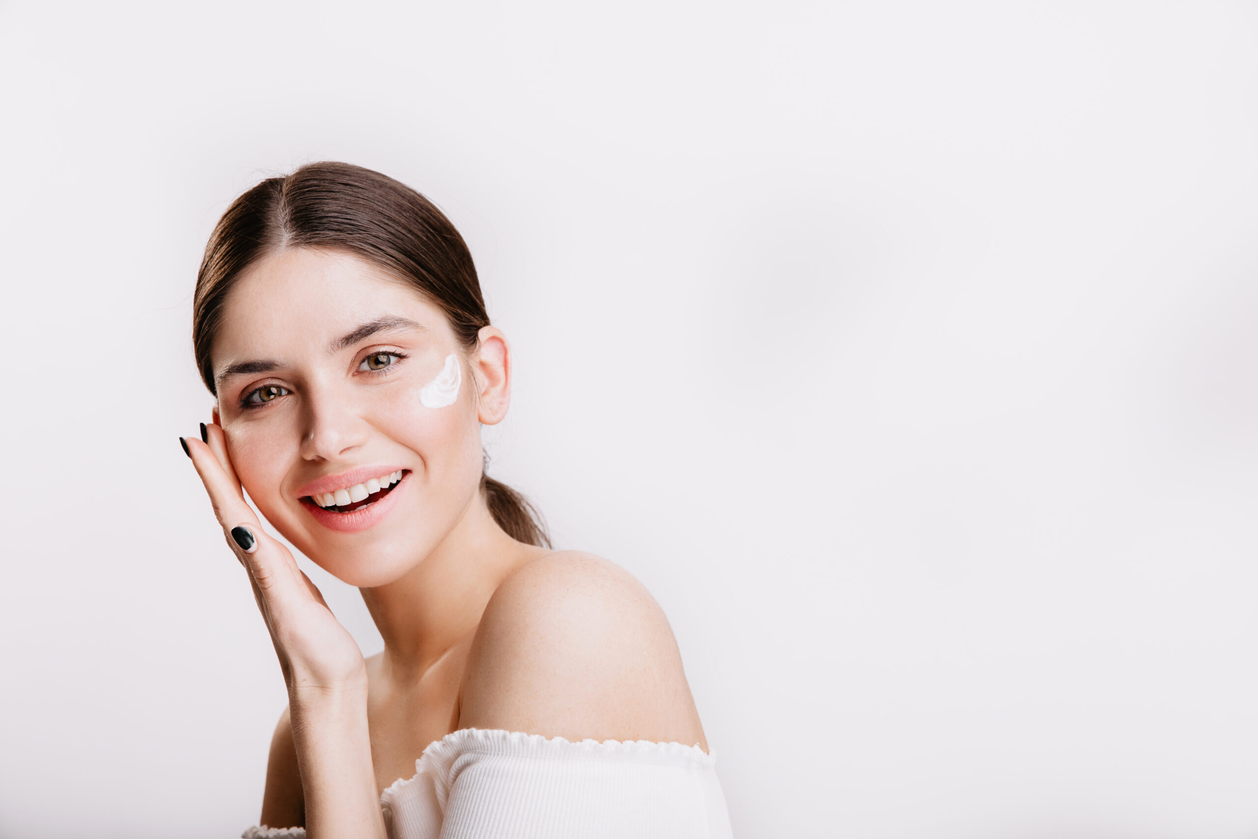 Girl touches moisturized skin and smiles. Portrait of model with cream on face on isolated background.